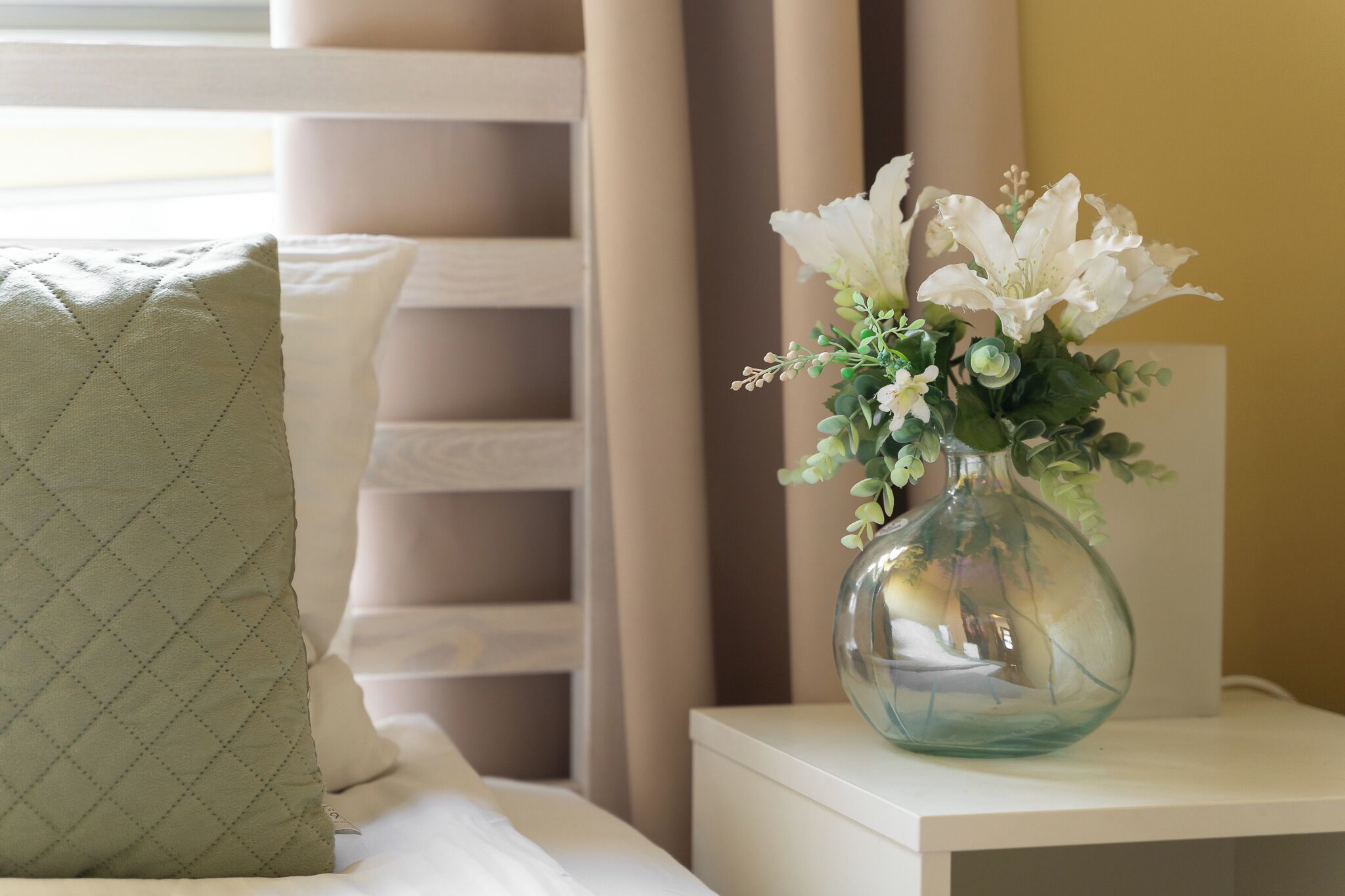 A neatly made bed with a green quilted pillow and a clear glass vase with white flowers on a bedside table.