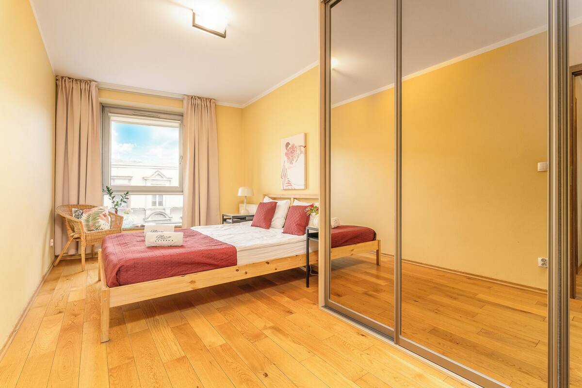 A bedroom with a wooden bed, a window with curtains, a bedside table, and a mirror.