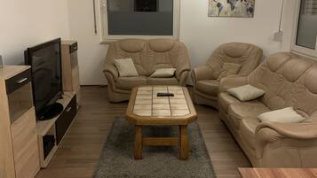 Ein Wohnzimmer mit Couchtisch aus Holz, zwei beigefarbenen Sofas, einem Fernseher und einer Weltkarte als Kunstwerk.