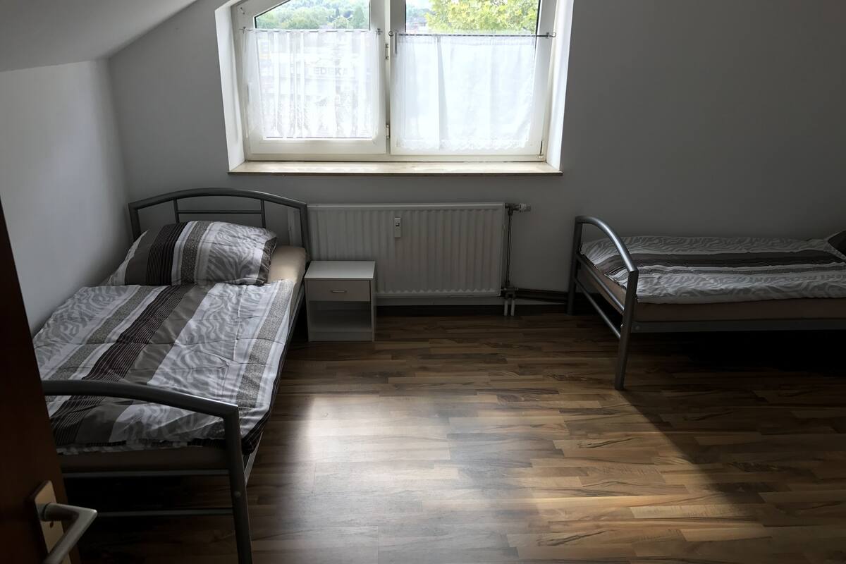 A dormitory room with two single beds, a small bedside table, a radiator, and a window with curtains.