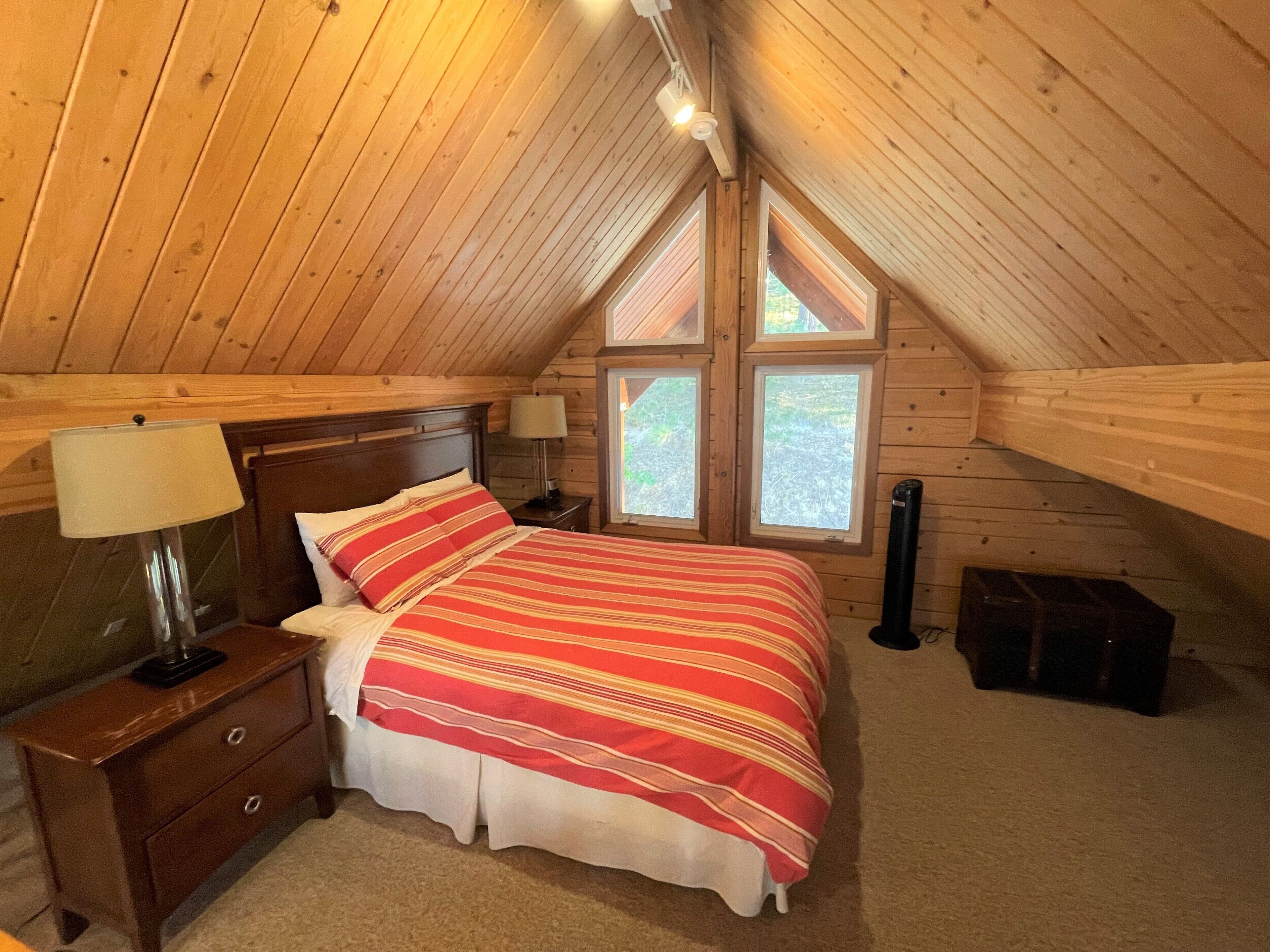 A cozy attic bedroom with a wooden sloped ceiling, a bed with striped bedding, two bedside lamps, and a window with a view of greenery.