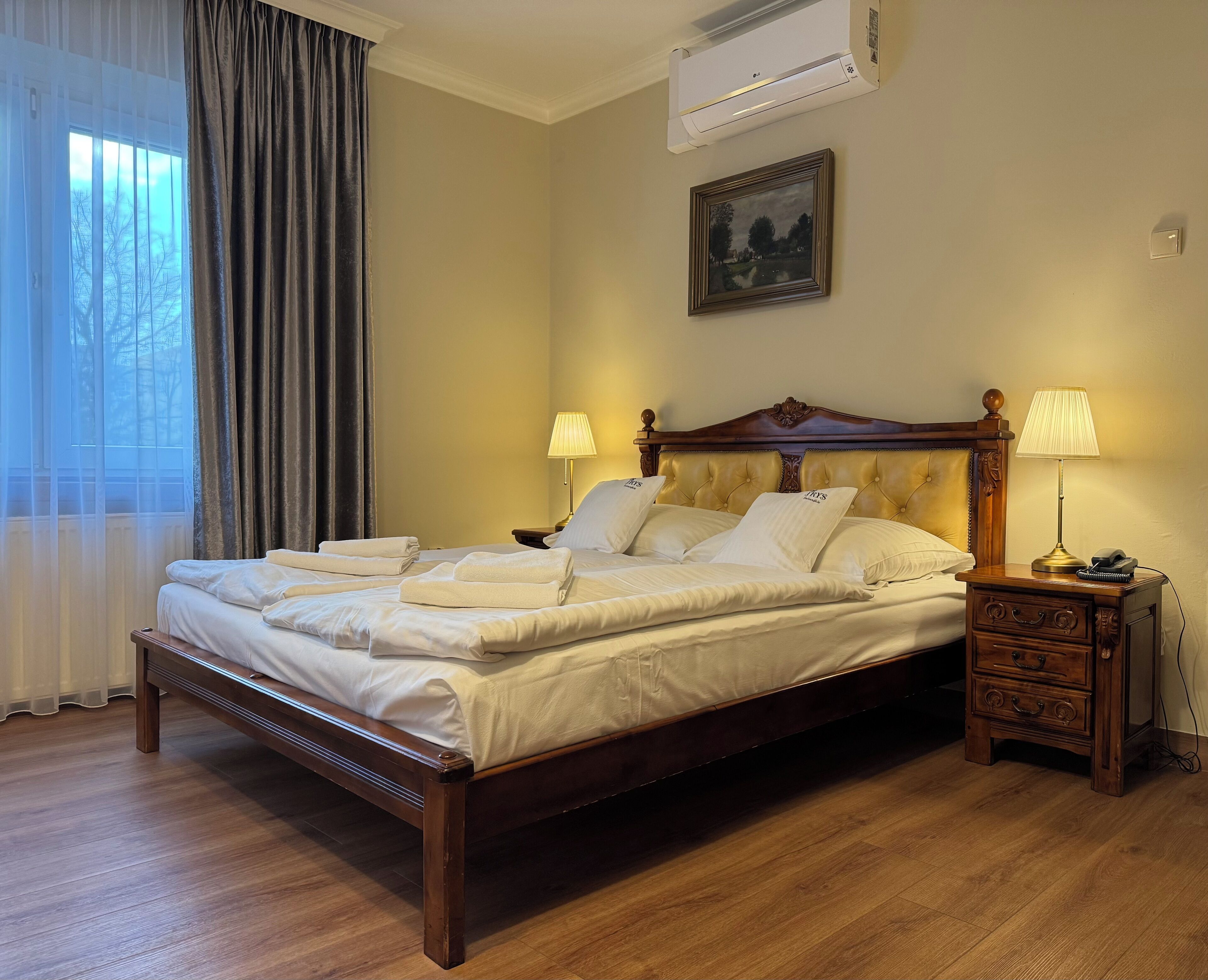 A neatly made bed with white linens and towels, a wooden headboard, a bedside table with a lamp, and a painting on the wall.