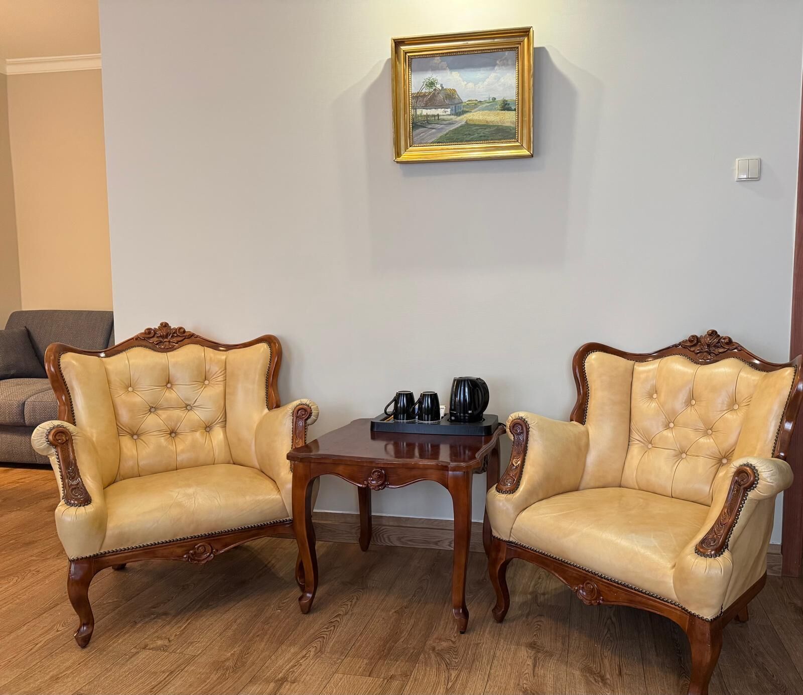Deux fauteuils à l’accoudoir, dotés d’un revêtement capitonné et de cadres en bois, une petite table en bois avec une machine à café, et un tableau représentant un paysage, encadré et accroché au mur.