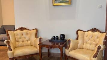Dos sillones con tapicería capitonada y armazones de madera, una mesita de madera con una cafetera, y un cuadro con paisaje en la pared.