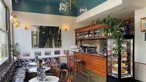 Interior de restaurante con zona de bar, mesas redondas y bancos acolchados, y una vitrina con postres.