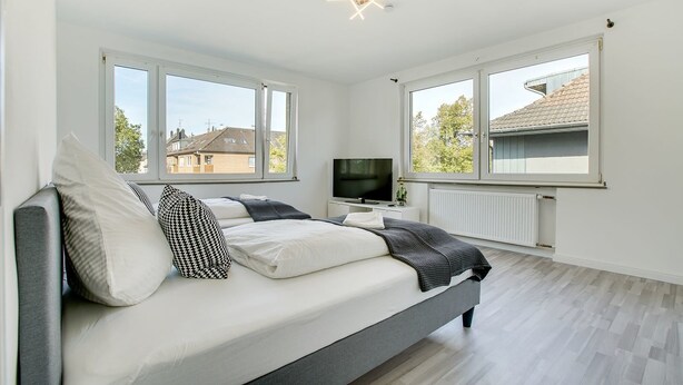A modern bedroom with a bed, a TV, and large windows. - Stay Here ND Apartments (Krefeld)