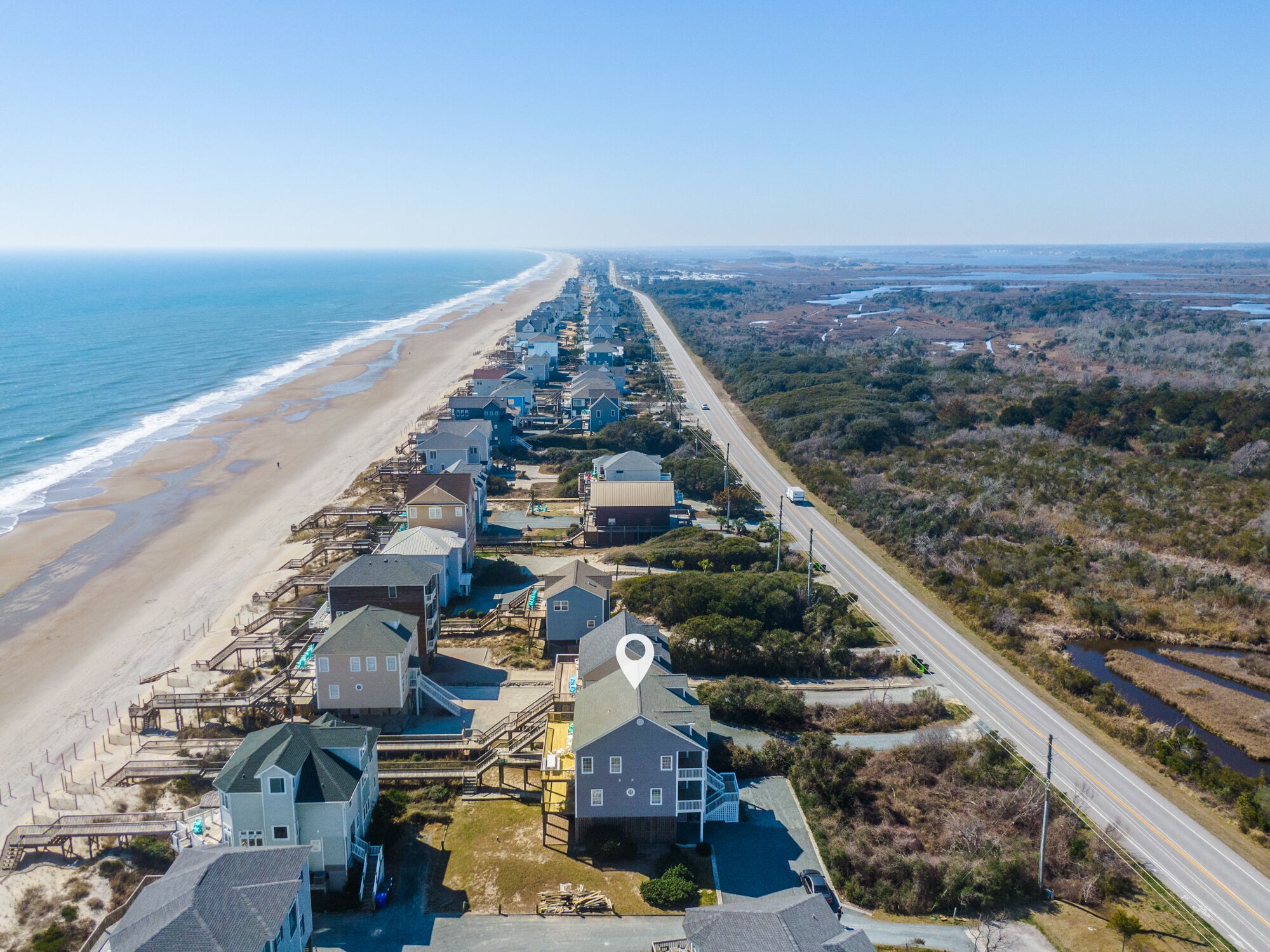 An aerial view of a coastal residential area with houses along a beachfront, a road parallel to the beach, and a body of water in the distance.
