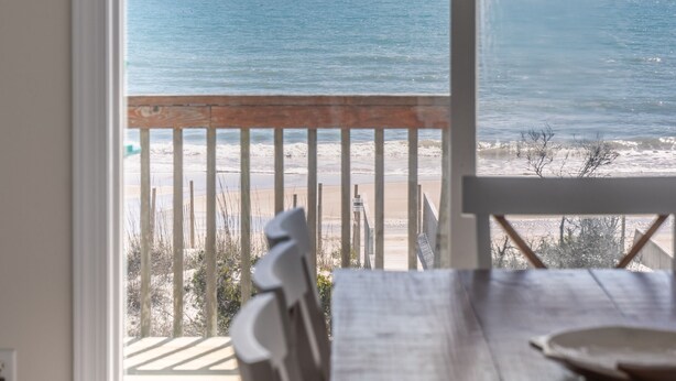 A view from a balcony overlooking the ocean, with a wooden table and chairs in the foreground. - LegaSea | Private Beach Access, Pool + Hot Tub! (North Topsail Beach)