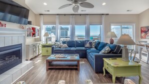 A spacious living room with a large blue sofa, a fireplace, a ceiling fan, and a view of the ocean. - LegaSea | Private Beach Access, Pool + Hot Tub! (North Topsail Beach)