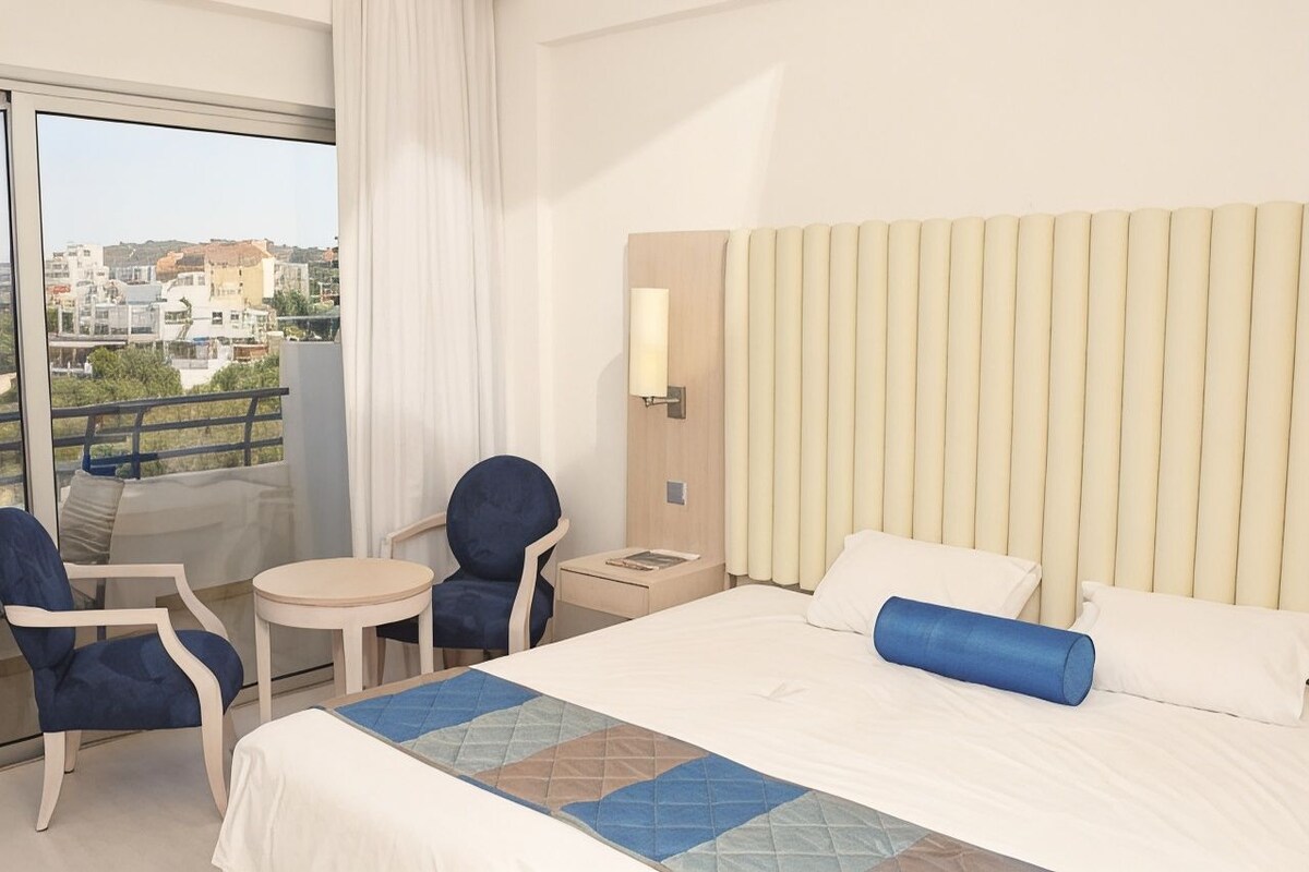 Quarto de hotel com uma cama grande, duas cadeiras, uma mesinha, um criado-mudo, um abajur e vista para a cidade através de uma janela ampla.
