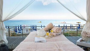 Een bed aan het strand met uitzicht op zee, handdoeken en een dienblad met een schaal en bloemen.