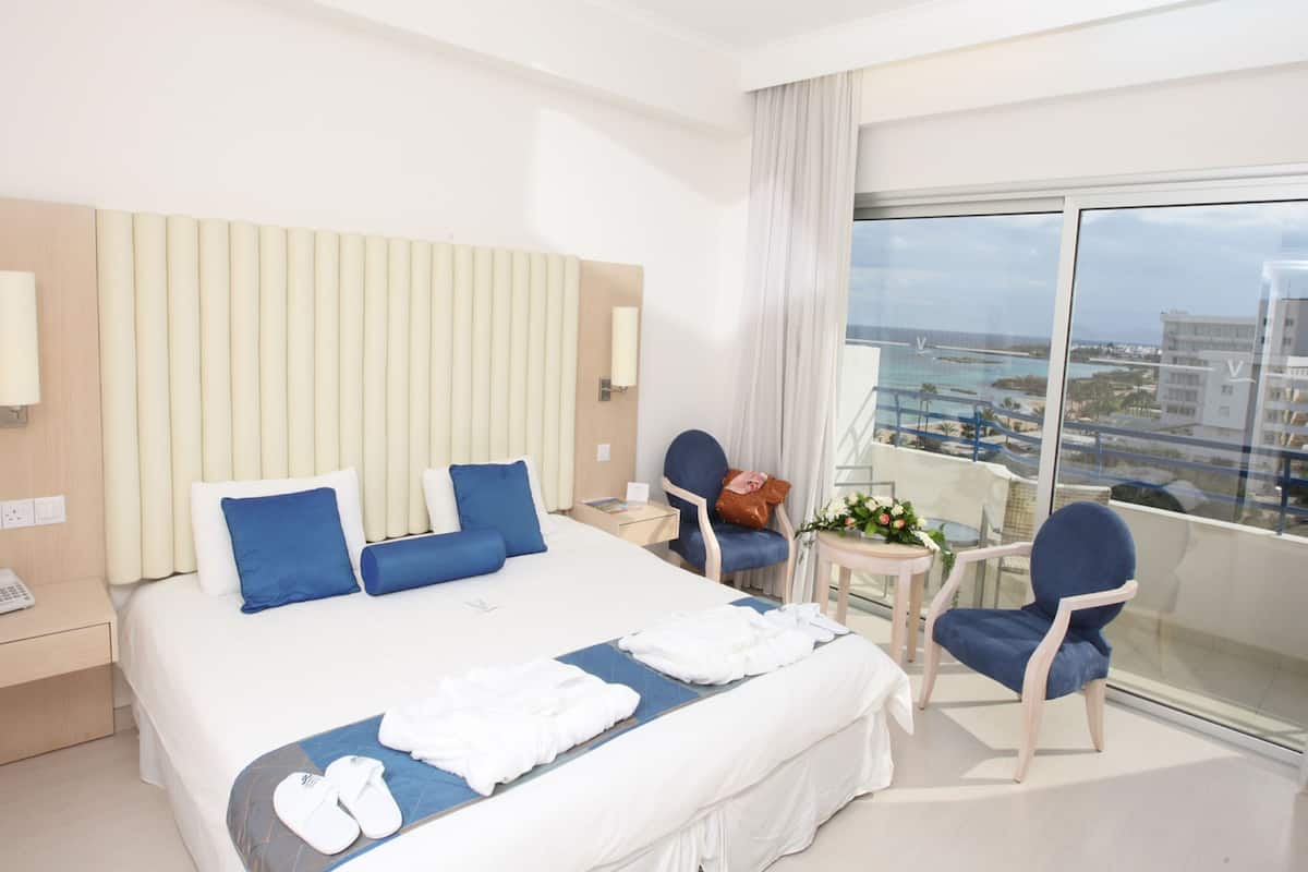 Habitación de hotel con una cama grande, una mesita con un florero, dos sillas y un balcón con vistas a la ciudad.