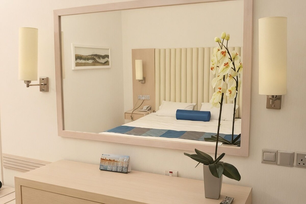 Quarto de hotel com um espelho grande, uma cama com travesseiro azul, mesa de cabeceira com um livro e um vaso de plantas.