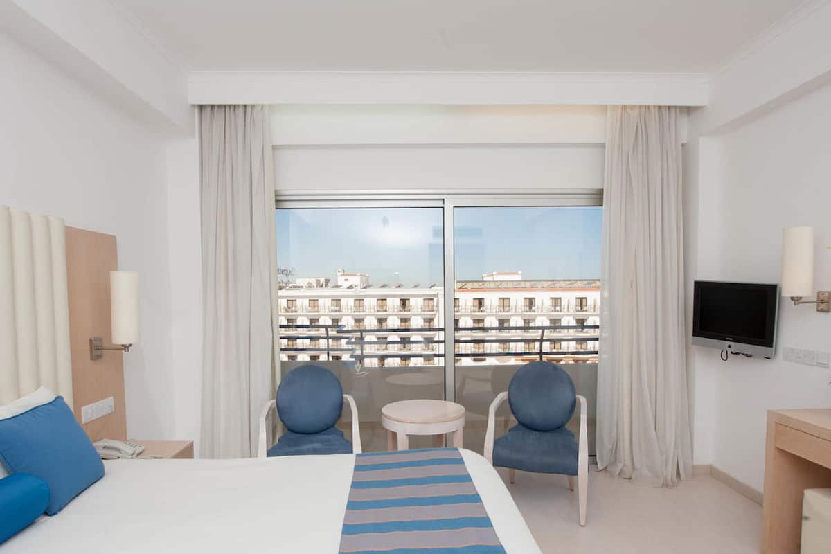 Habitación de hotel con cama, dos sillas, una mesita, televisor y balcón con vista a la ciudad.