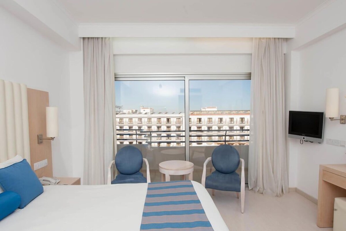 Quarto de hotel com cama, duas cadeiras, uma mesa pequena, televisão e varanda vista da cidade.