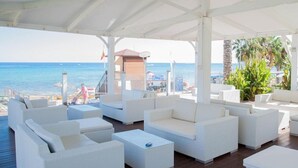 Een overdekt terras met witte rieten meubels, met uitzicht op een strand en de zee.