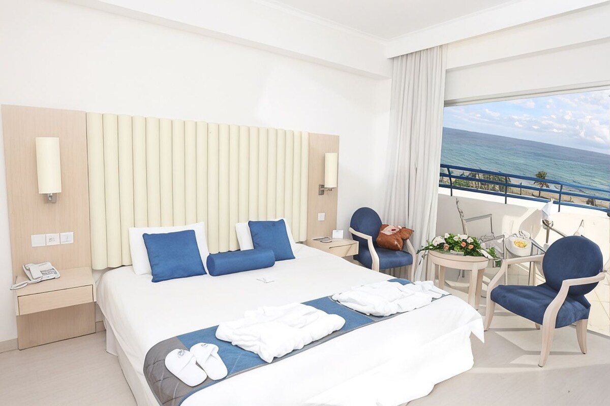 Quarto de hotel com cama, mesas de cabeceira, uma cadeira, uma pequena mesa com arranjo de flores e vista para o oceano.