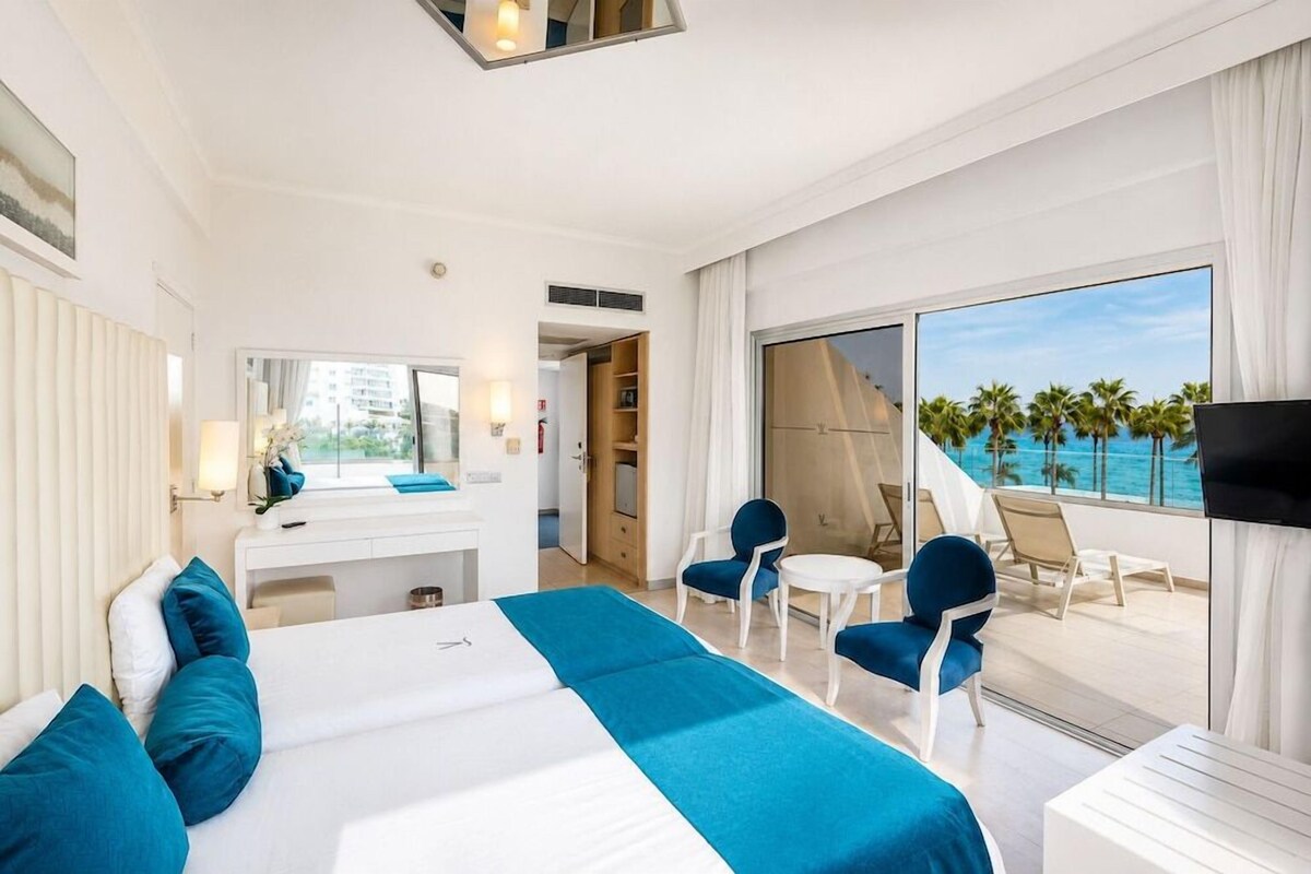 Quarto de hotel com cama, escrivaninha, cadeira, mesa pequena e varanda com vista para palmeiras e o oceano.