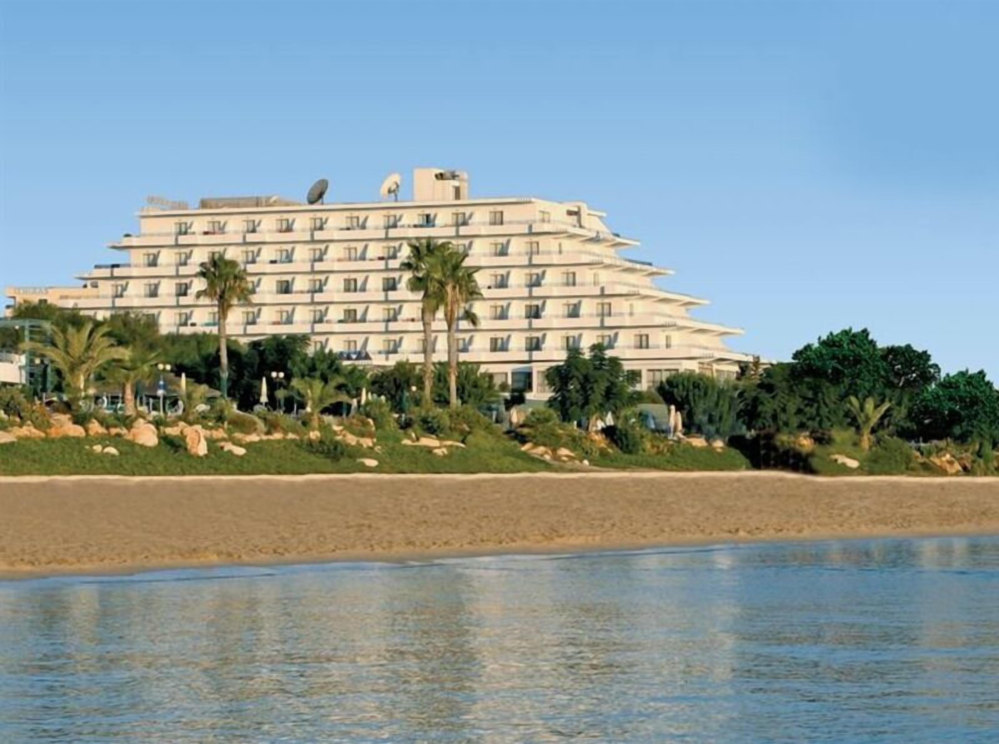 A multi-story hotel with a flat roof, located near a beach with palm trees and a sandy shoreline.