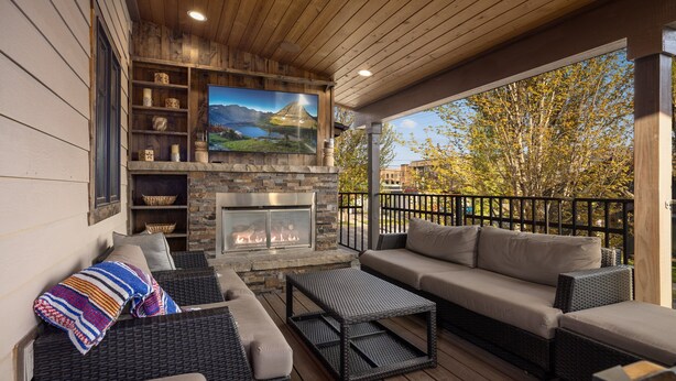 A covered outdoor seating area with wicker furniture, a stone fireplace, and a television mounted above it.
