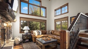 A spacious living room with a stone fireplace, large windows, a leather sofa, and a wooden staircase.