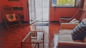 A room with a red wall, a window with a view of trees, a chair, a table, and a lamp. - WHITE & BLUE HOUSE (Chiavari)