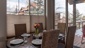 Un coin repas avec une table dressée pour quatre personnes, de grandes fenêtres, des chaises en osier et un vase rempli de fleurs.