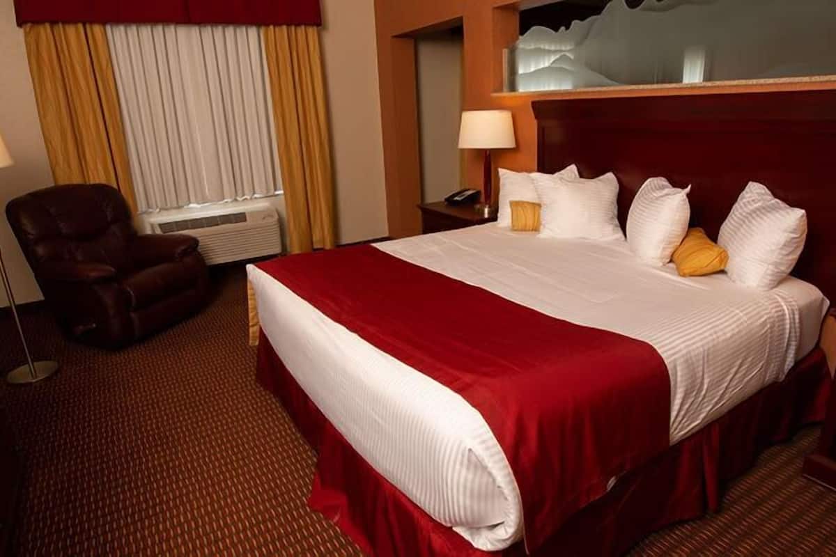Una habitación de hotel con una cama grande, un sillón de cuero, una mesita de noche con lámpara y una ventana con cortinas.