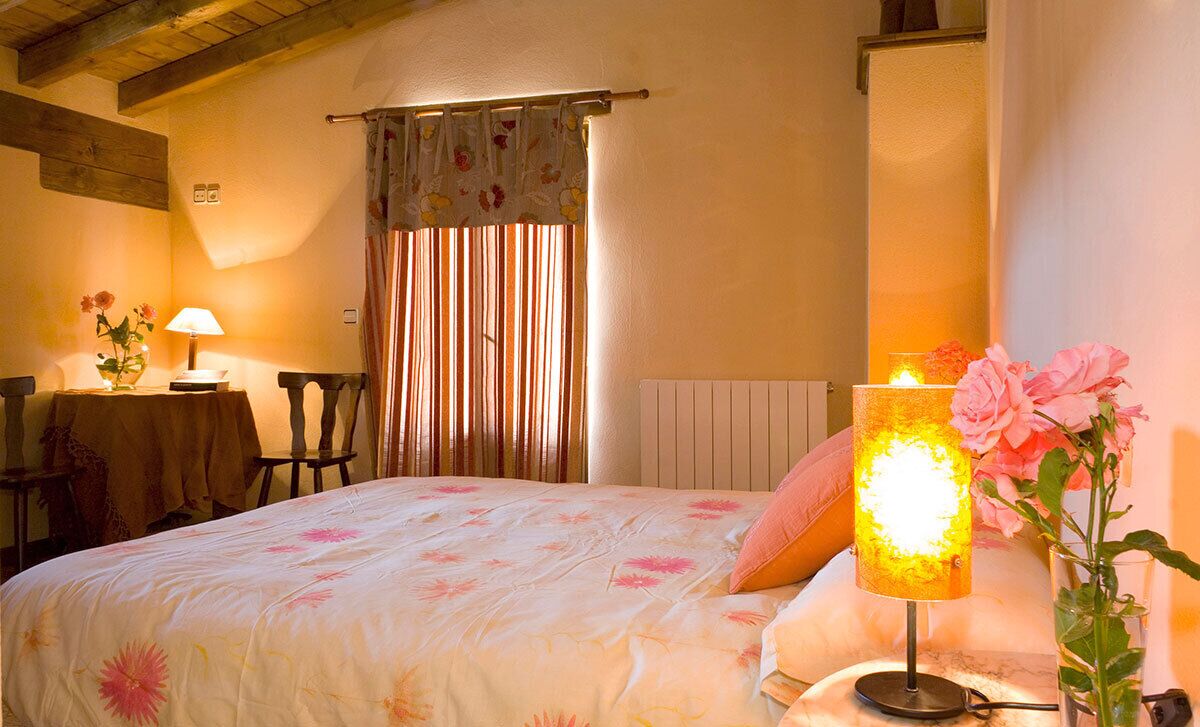Un dormitorio acogedor con una cama, una mesita de noche con lámpara, una silla y un ventanal con cortinas.