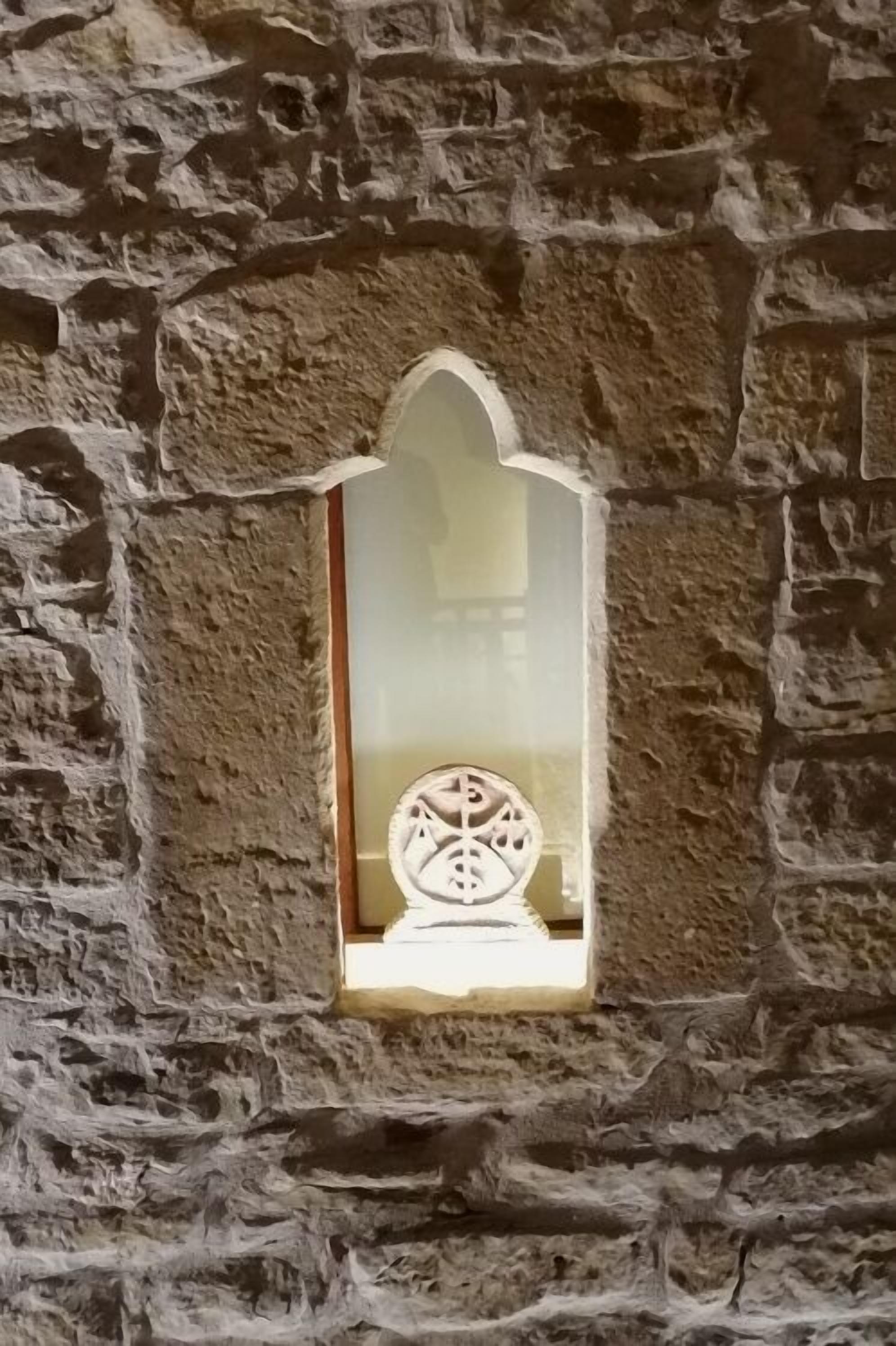 A stone wall with a narrow arched window featuring a decorative metal piece.