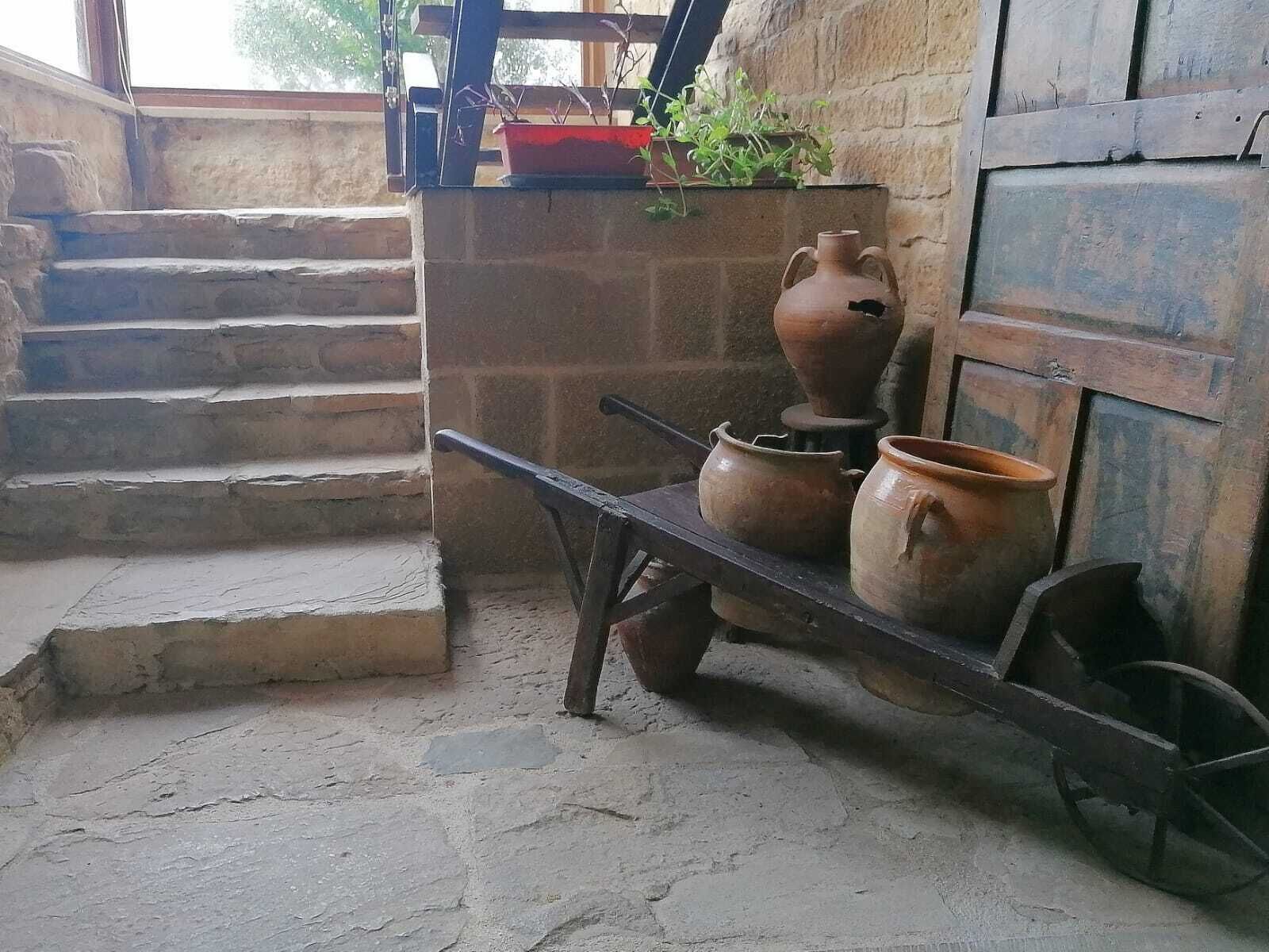 Un interior rústico con escalones de piedra que conducen a una puerta de madera, un carro con macetas de barro y una ventana con plantas.