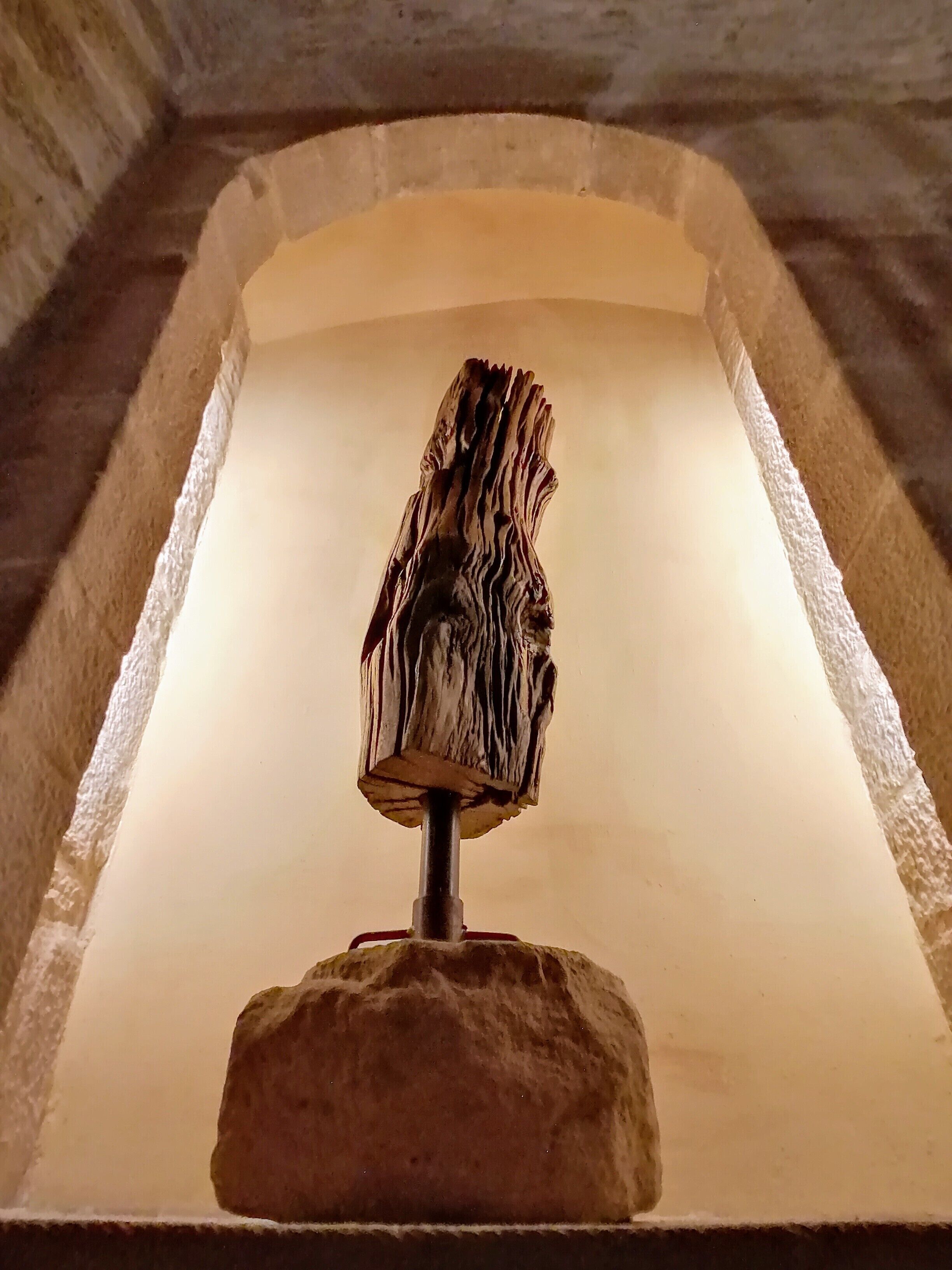 Estrutura de pedra com uma figura de madeira entalhada, possivelmente um artefato histórico, exposta em um ambiente com pouca luz e teto abobadado.