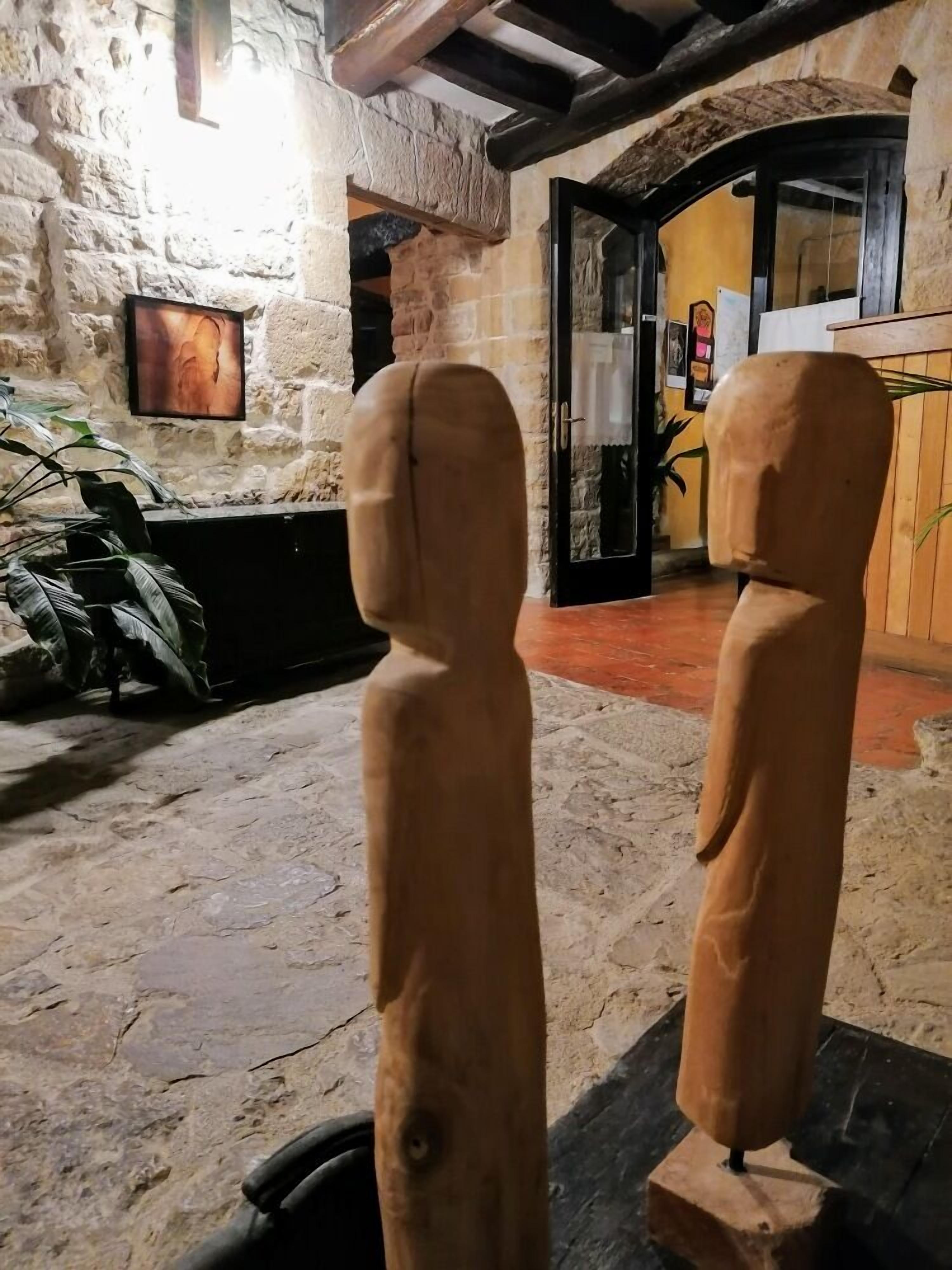 Two wooden sculptures in a stone-walled room with a television on the wall and a door leading to another room.