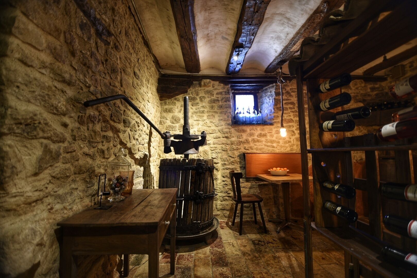 A rustic cellar with stone walls, a wooden table, a chair, a wine rack, and a window with a view.
