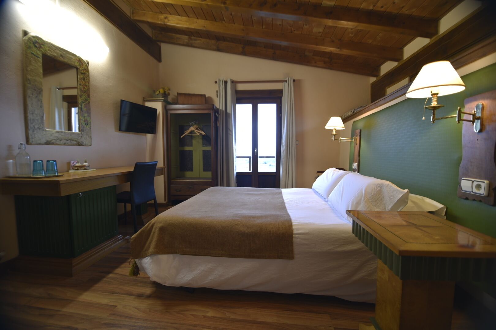 A hotel room with a bed, bedside table, mirror, TV, and a view of the outside through a window.