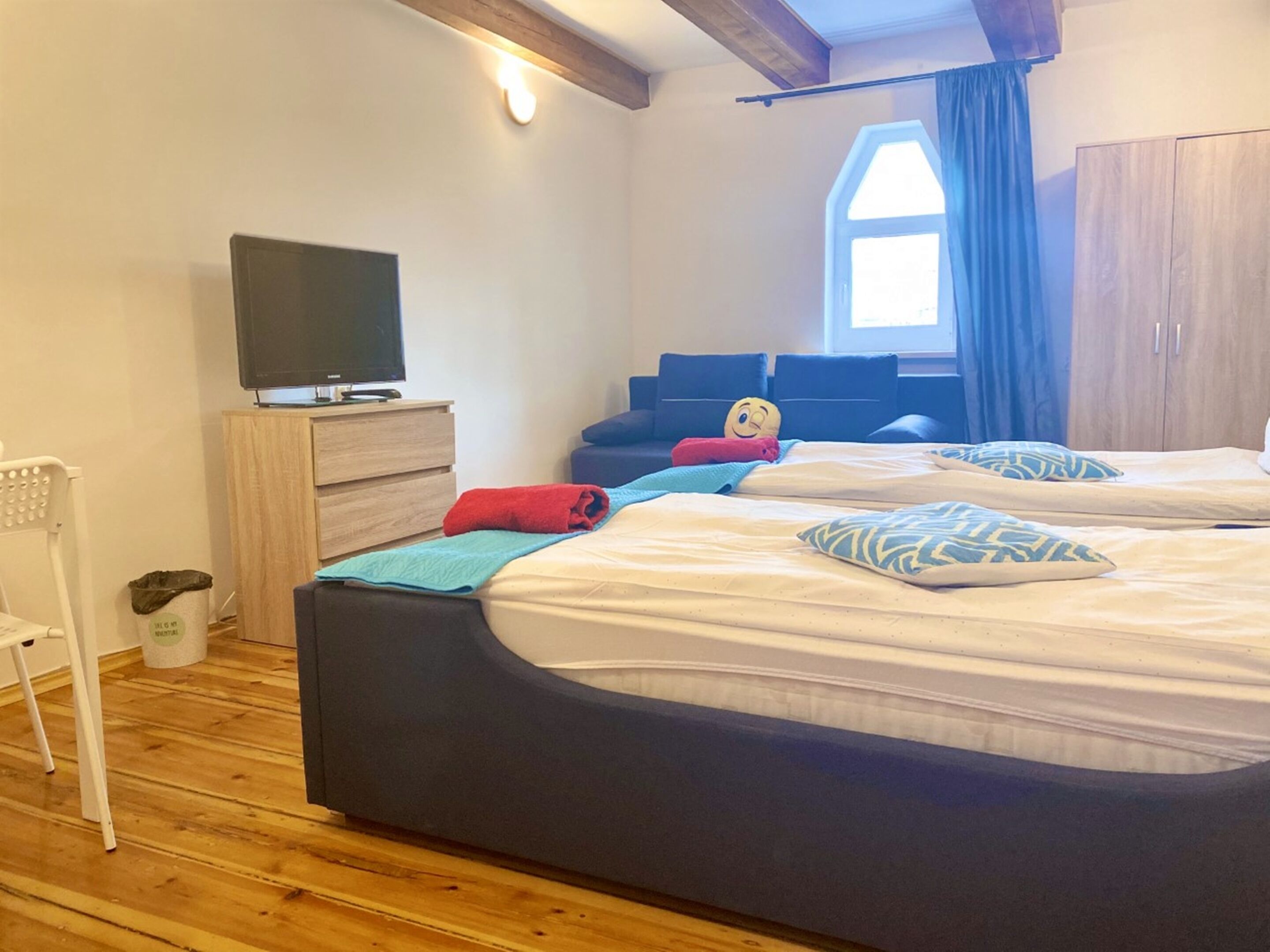 Un dormitorio con dos camas, un televisor sobre un aparador de madera, una ventana con cortinas y suelo de madera.