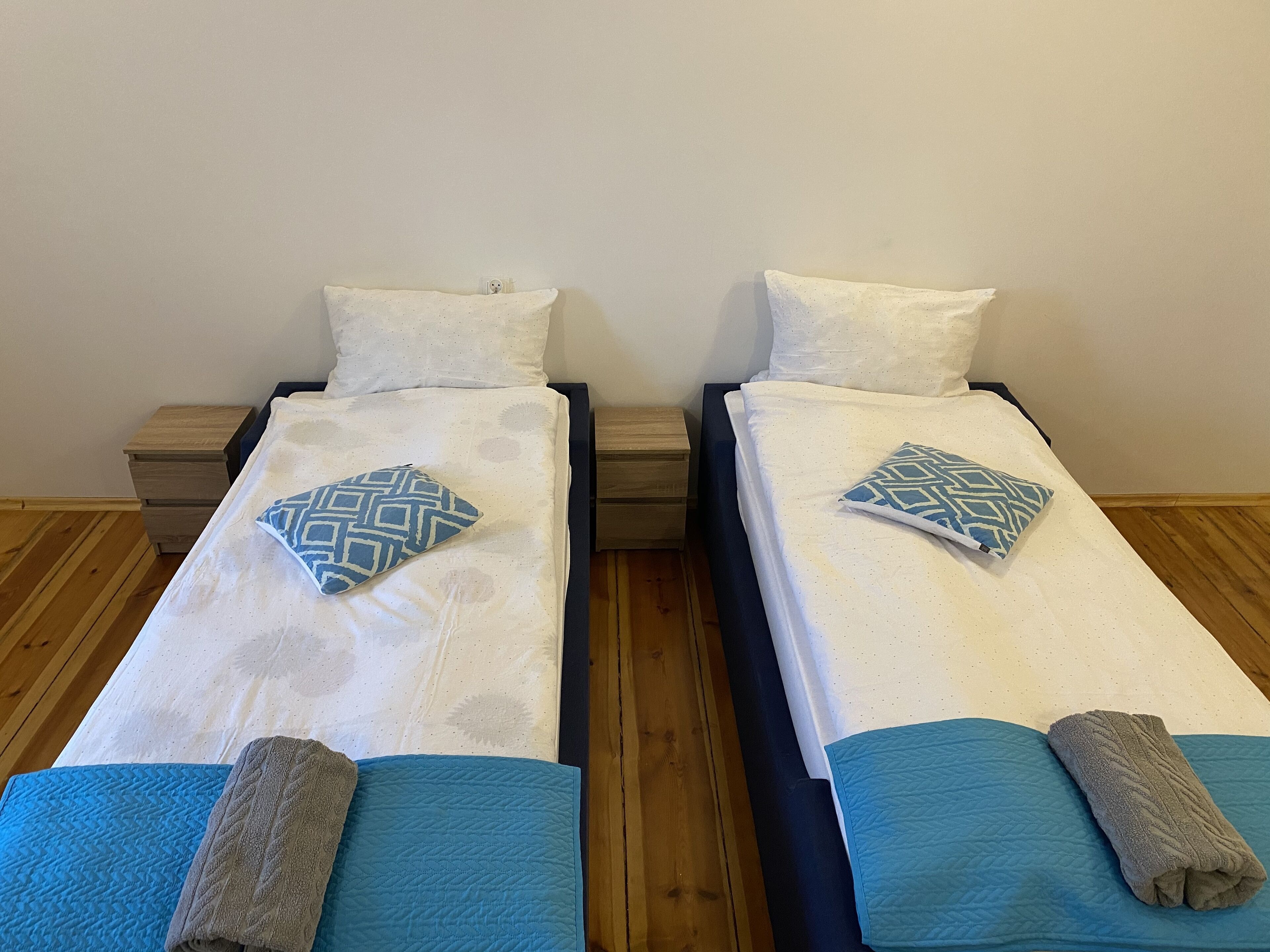 Duas camas de solteiro com almofadas estampadas e toalhas dobradas, mesinhas de cabeceira de madeira e uma parede lisa.