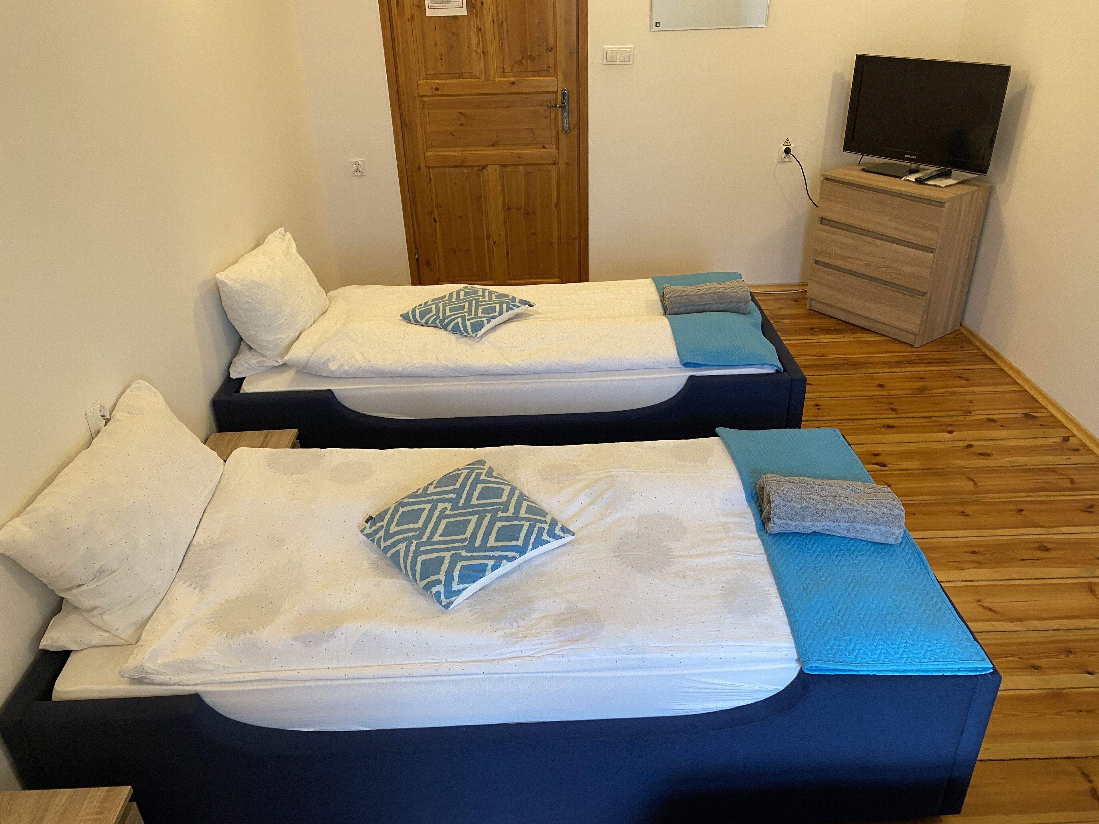 Two single beds with patterned pillows, a wooden dresser, and a television in a room with wooden flooring.