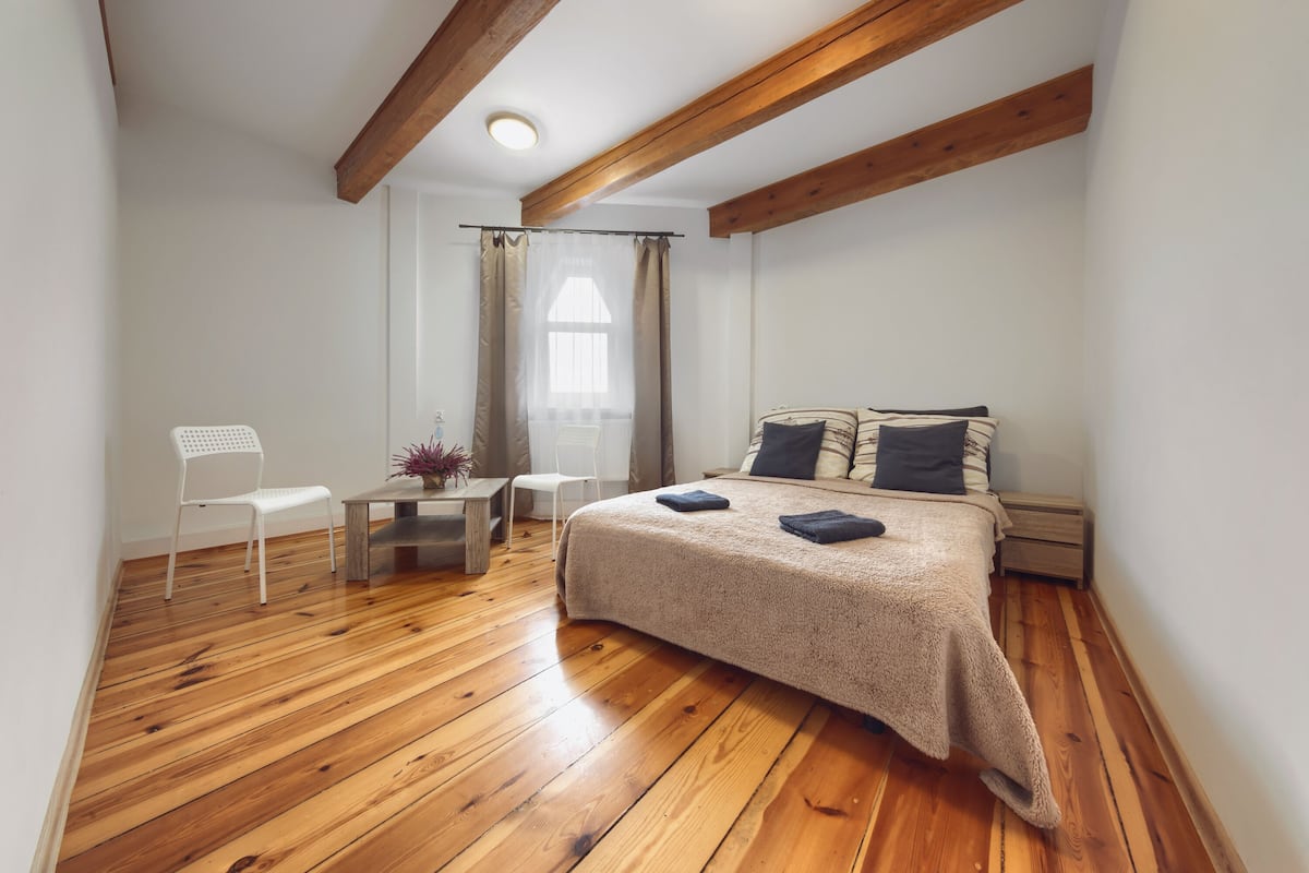 A bedroom with a wooden floor, a bed with a beige blanket, a white chair, a small table with a vase, and a window with curtains.