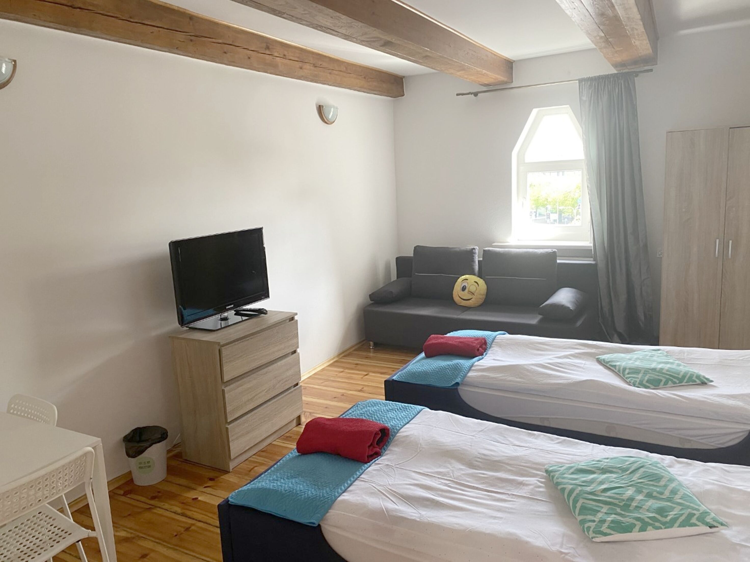 Ett sovrum med två sängar, en tv på en byrå, en soffa med en kudde som har ett leende på sig och ett fönster med gardiner.