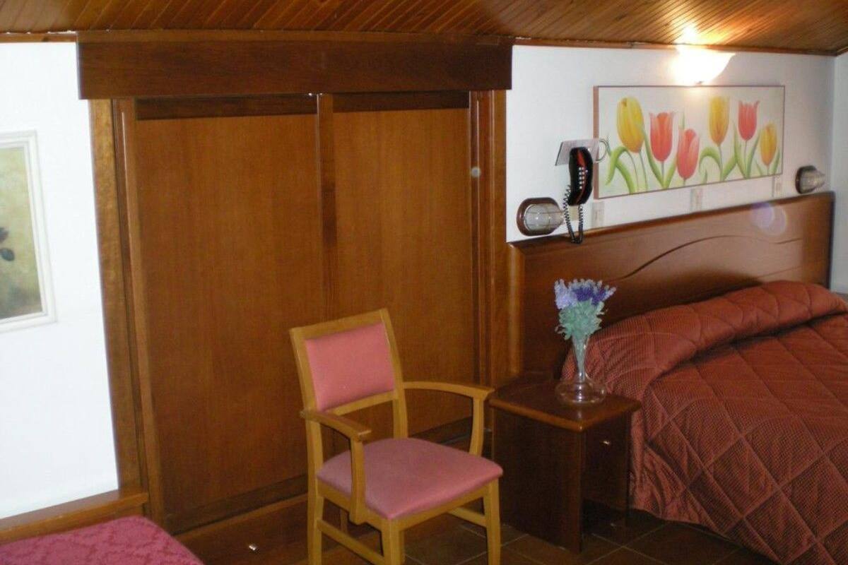 A bedroom with a wooden ceiling, a bed with a red quilt, a wooden chair, a bedside table with a vase, and a wall-mounted telephone.