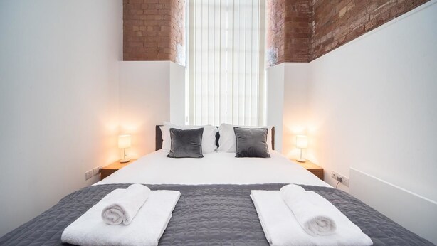 A hotel room with a bed, two bedside lamps, a window with blinds, and a brick wall.