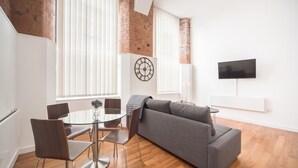 A modern living room with a gray sofa, a glass dining table with chairs, a brick wall, and a flat-screen TV mounted on the wall.