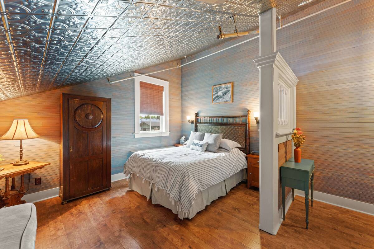 Un dormitorio con piso de madera, una cama con cabecera, una ventana con persianas, una puerta y una lámpara sobre una mesita.