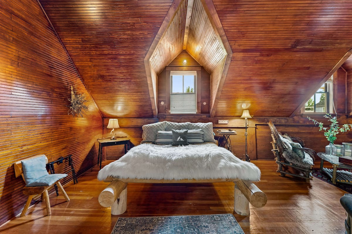 Un dormitorio acogedor en el ático, con techo inclinado de madera, una cama grande con ropa blanca, una mecedora y un ventanal pequeño.