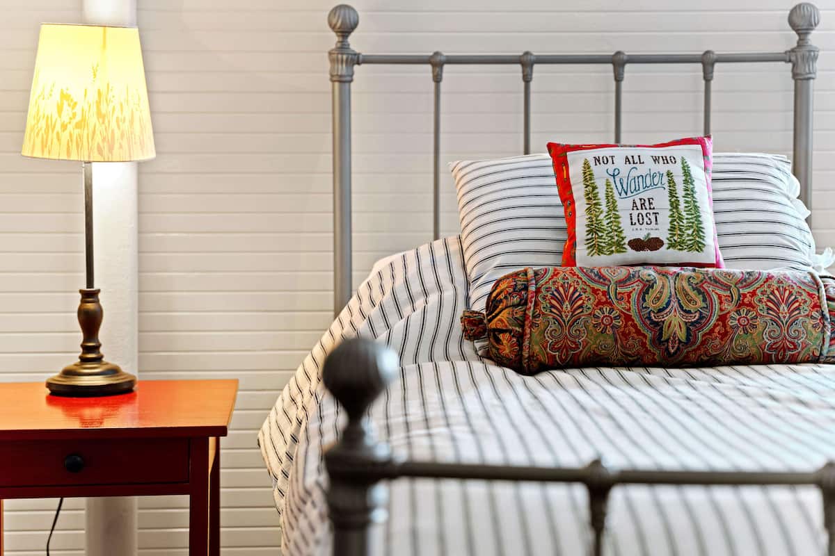 Una cama bien tendida con ropa de cama a rayas y almohadones decorativos, una lámpara de noche y un cabecero de metal.