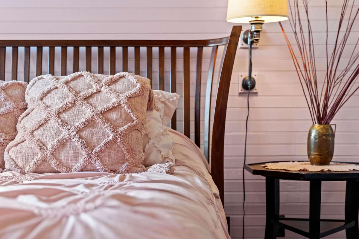 Una cama con manta y almohadas con textura, un cabecero de madera, una lámpara fijada a la pared y una pequeña mesa con un florero.