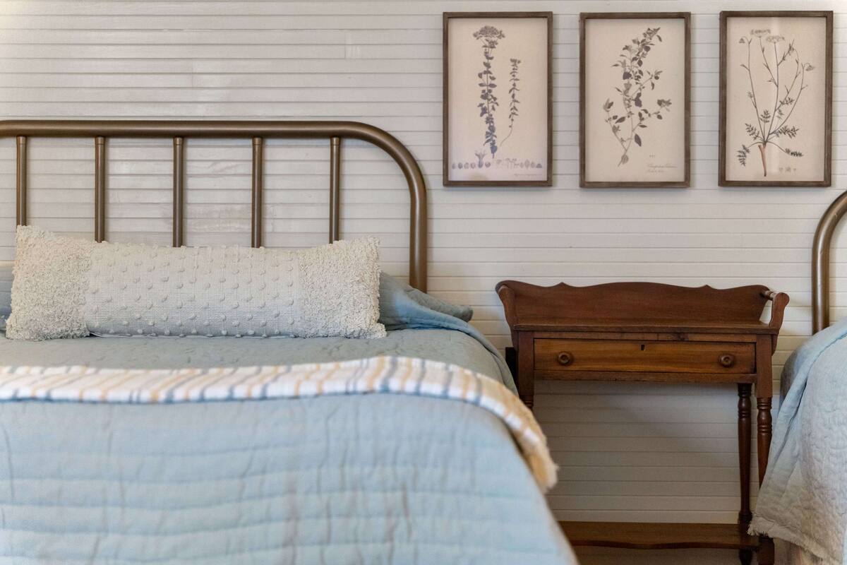 Un dormitorio con estructura de cama metálica, mesita de noche de madera y estante montado en la pared. En la pared hay ilustraciones botánicas enmarcadas.