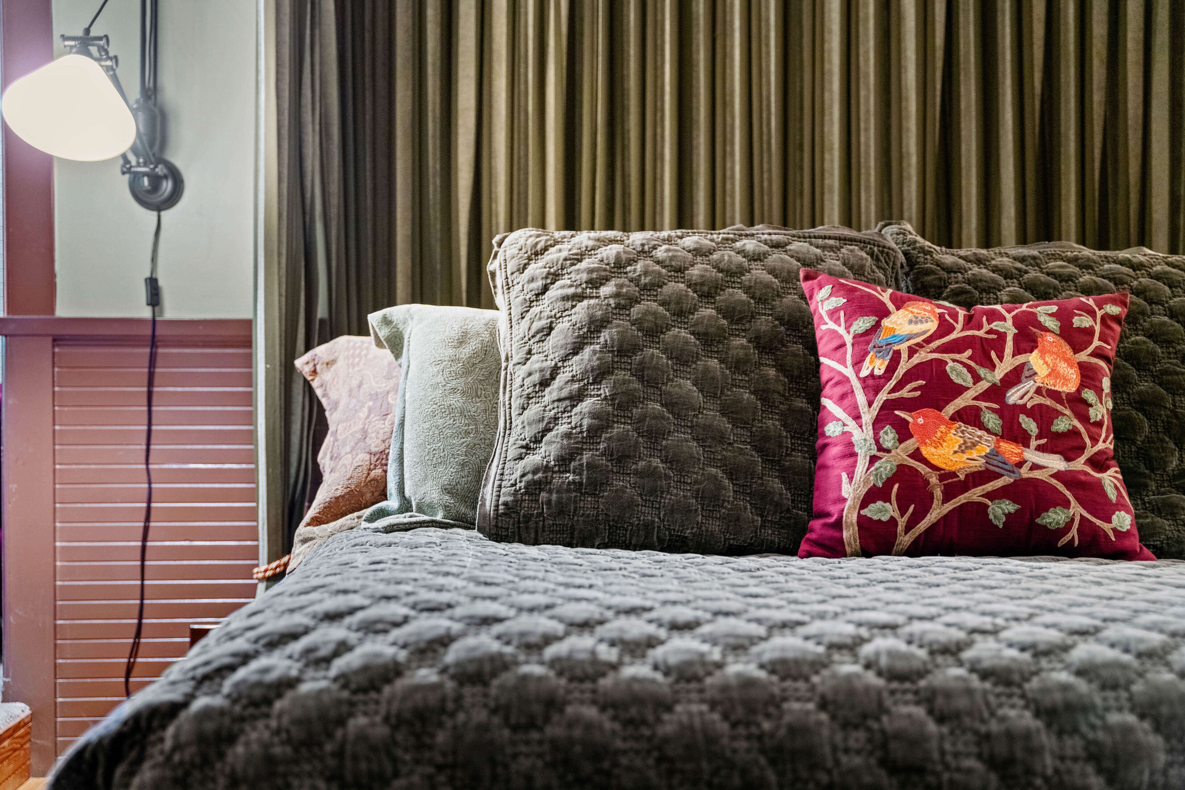 A bed with multiple pillows, a wall-mounted lamp, and a window with blinds.
