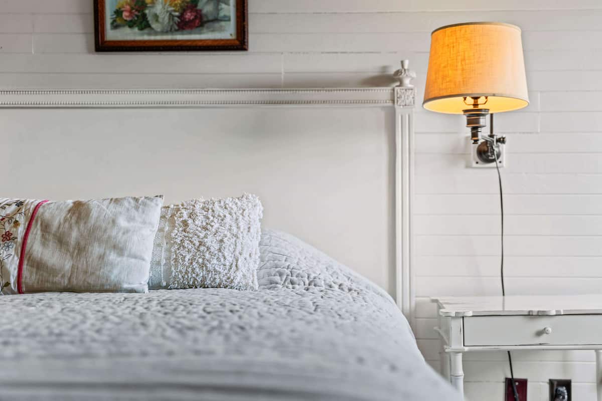 Una cama bien tendida con un cojín de encaje y una manta texturizada, una lámpara de pared y una mesita de noche blanca con un cajón.